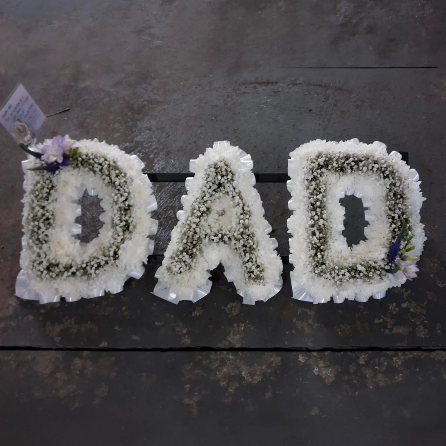 Funeral Flower Letters – White Base by ReCreation Flowers spell DAD in white blooms, with purple accents on the first and last letters for a striking custom tribute, displayed on a dark surface.