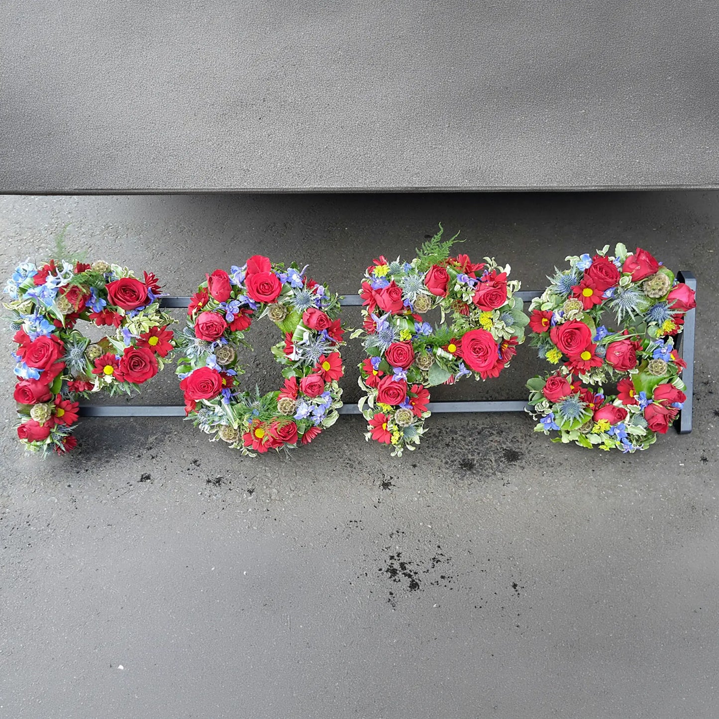 ReCreation Flowers’ Funeral Flower Letters – Open Loose Style spells “POPS” with red, purple, yellow, and blue blooms arranged on a gray surface.