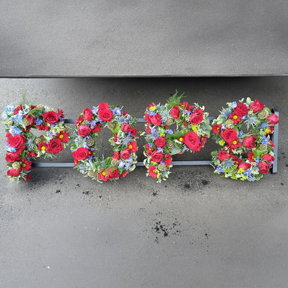 ReCreation Flowers’ Funeral Flower Letters – Open Loose Style spells “POPS” with red, purple, yellow, and blue blooms arranged on a gray surface.