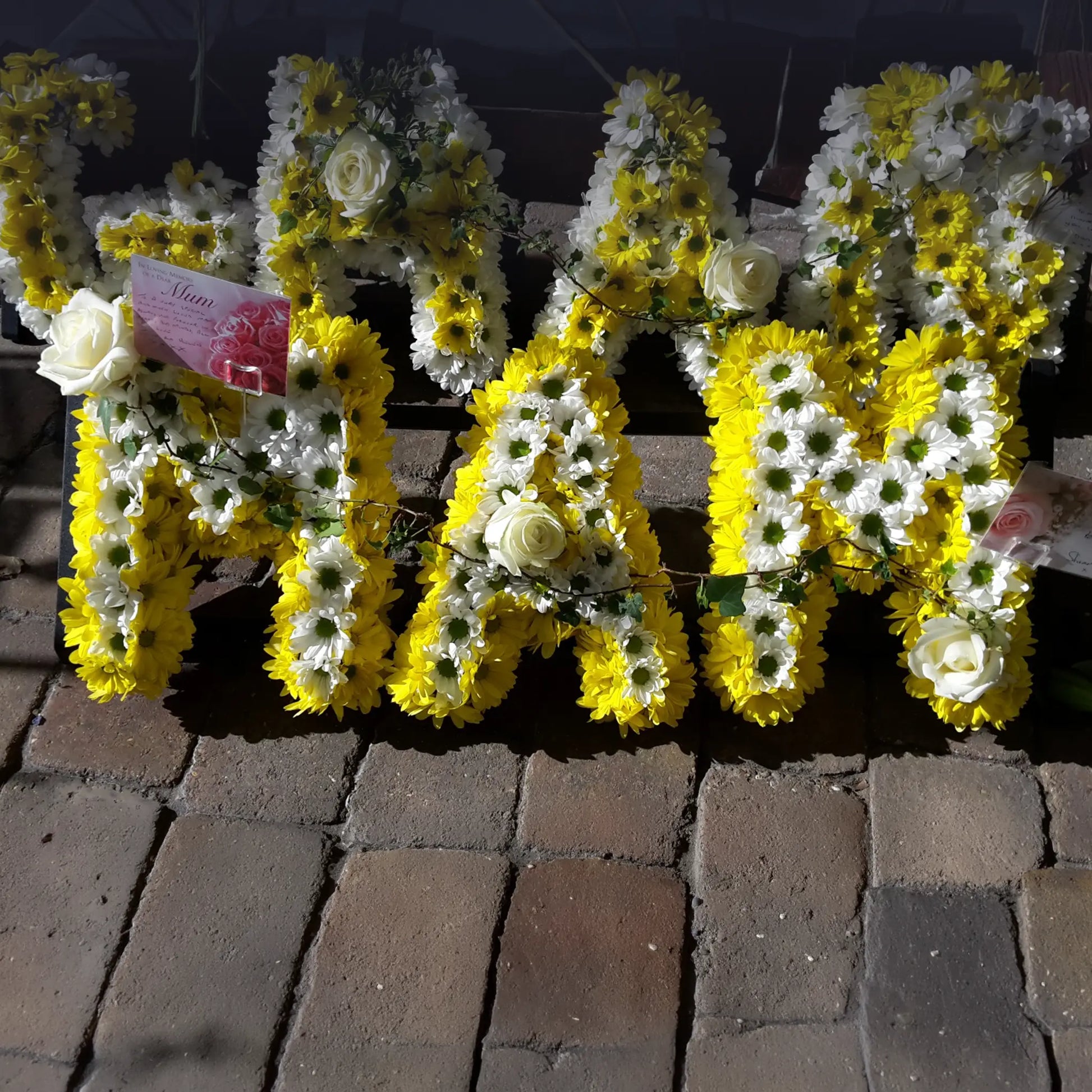 The Funeral Flower Letters – White Base by ReCreation Flowers feature custom floral letters spelling MAM with white and yellow flowers and white roses, displayed on stone pavement with attached cards.