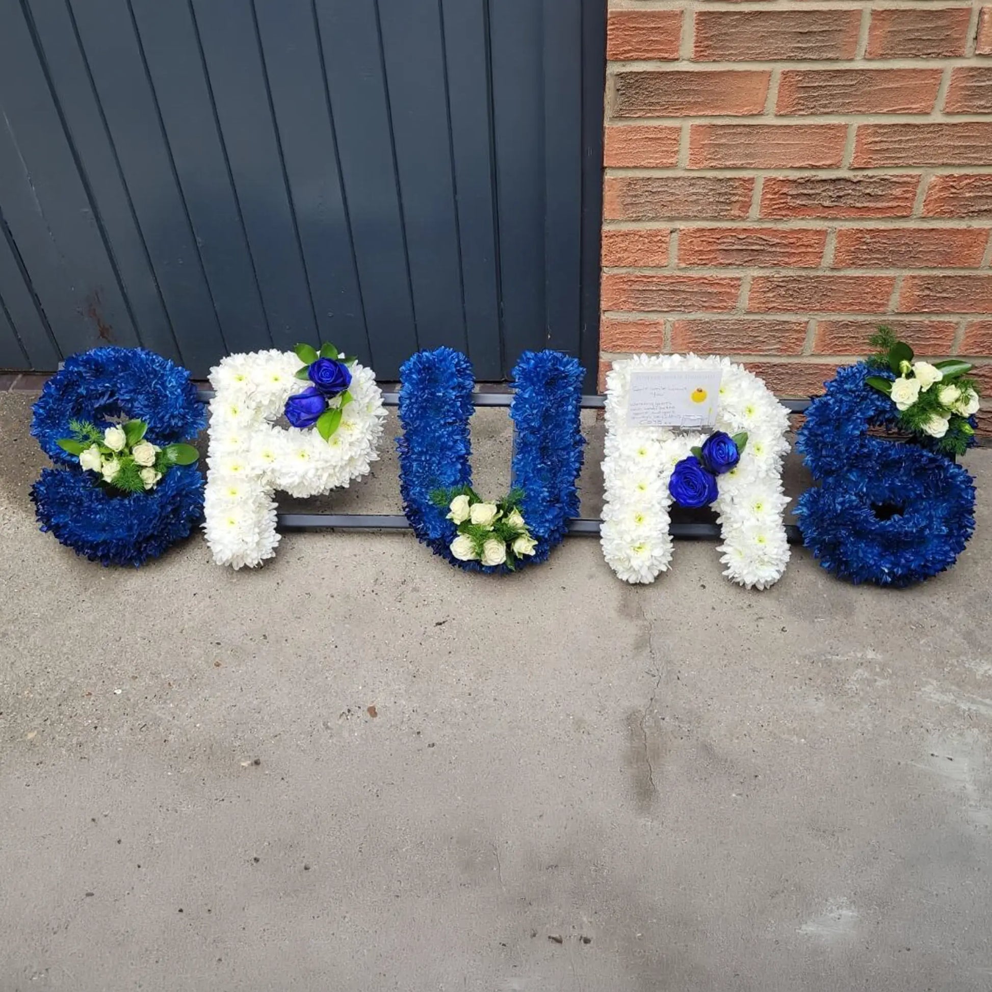 ReCreation Flowers’ Funeral Flower Letters – White Base spells out "SPURS" in blue and white, featuring small attached bouquets. The arrangement is displayed resting on the ground against a brick wall and gray garage door.