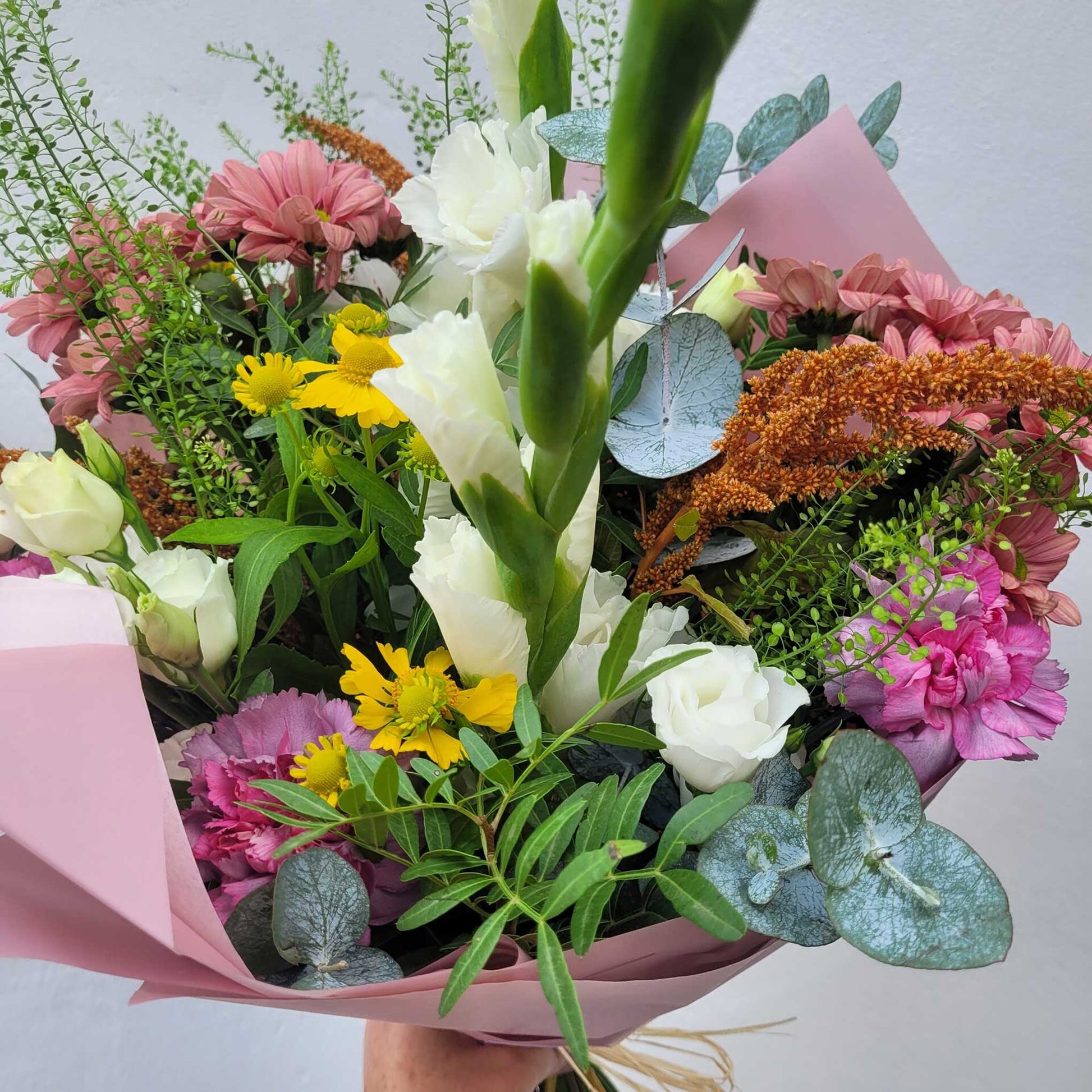 A hand holds the Florist Choice - Brights bouquet by ReCreation Flowers, showcasing vibrant seasonal blooms and lush foliage, wrapped in pink paper—crafted by a Leigh on Sea florist against a plain light background.