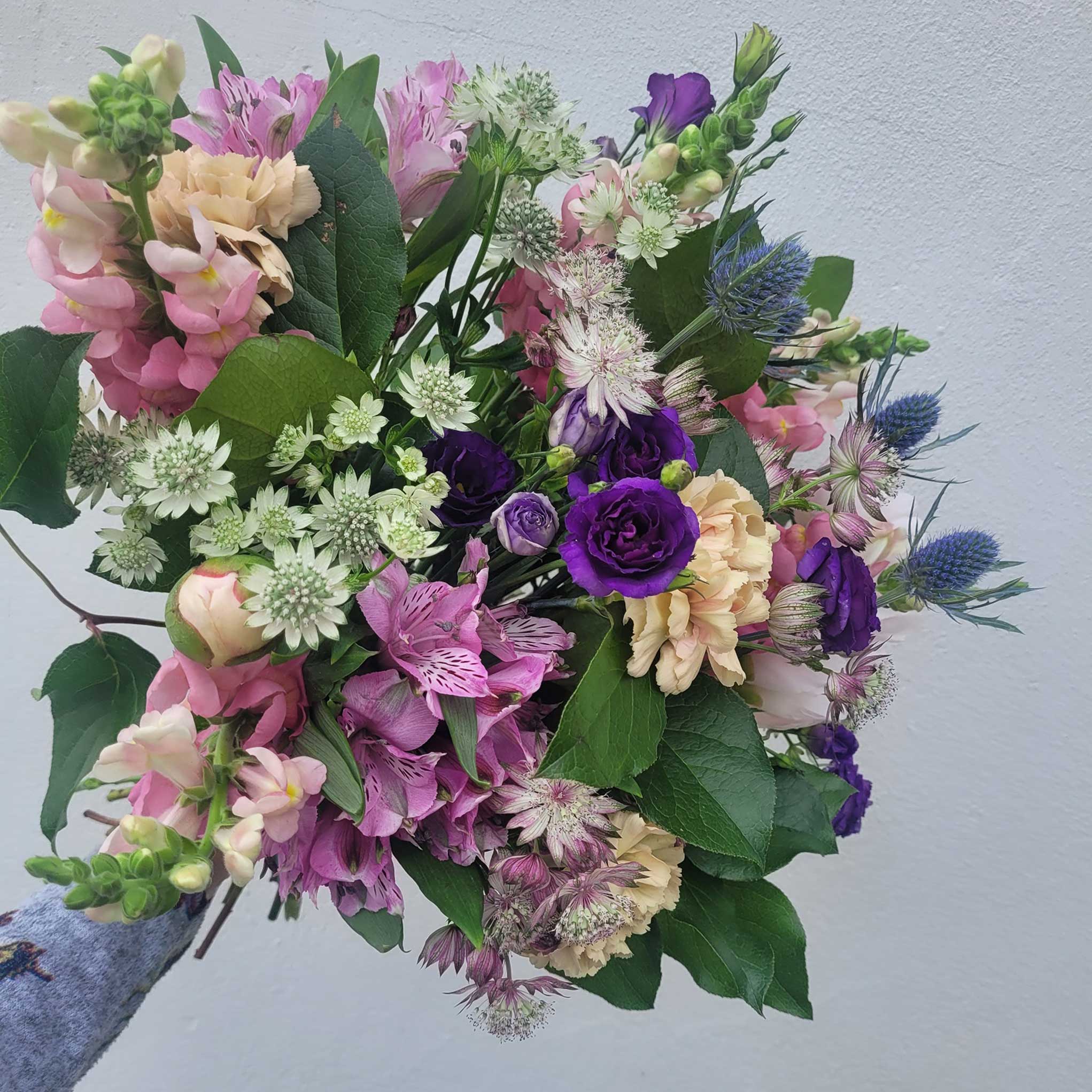 A hand holds the "Florist Choice - Pastel" bouquet by ReCreation Flowers, featuring pastel seasonal blooms and leafy stems against a light gray background—expertly arranged by your Leigh on Sea florist.