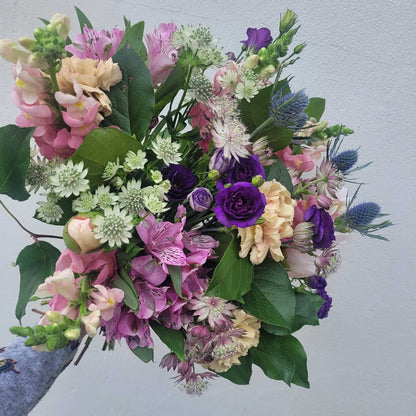 A hand holds the "Florist Choice - Pastel" bouquet by ReCreation Flowers, featuring pastel seasonal blooms and leafy stems against a light gray background—expertly arranged by your Leigh on Sea florist.