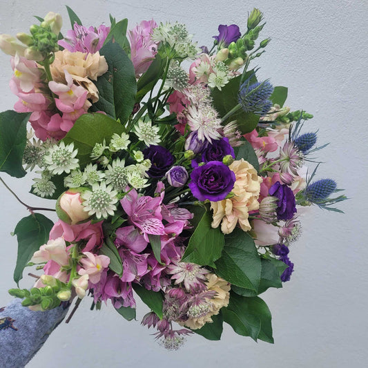 A hand holds the "Florist Choice - Pastel" bouquet by ReCreation Flowers, featuring pastel seasonal blooms and leafy stems against a light gray background—expertly arranged by your Leigh on Sea florist.