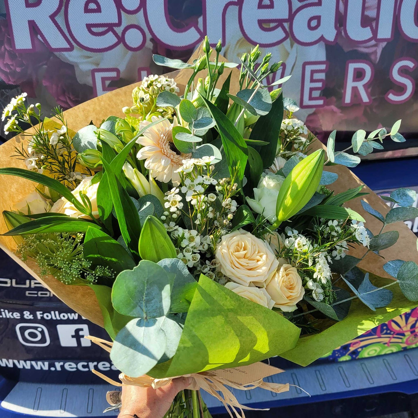 A hand holds the "Florist Choice - White & Green" bouquet by ReCreation Flowers, featuring cream and white seasonal blooms with greenery in brown paper. A banner with branding and social icons suggests Leigh on Sea florist charm.