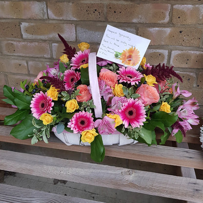 The Funeral Flower Basket by ReCreation Flowers features seasonal florist's choice pink, yellow, and peach blooms with mixed foliage. Presented with a condolence card, it is displayed on a wooden bench against a brick wall.