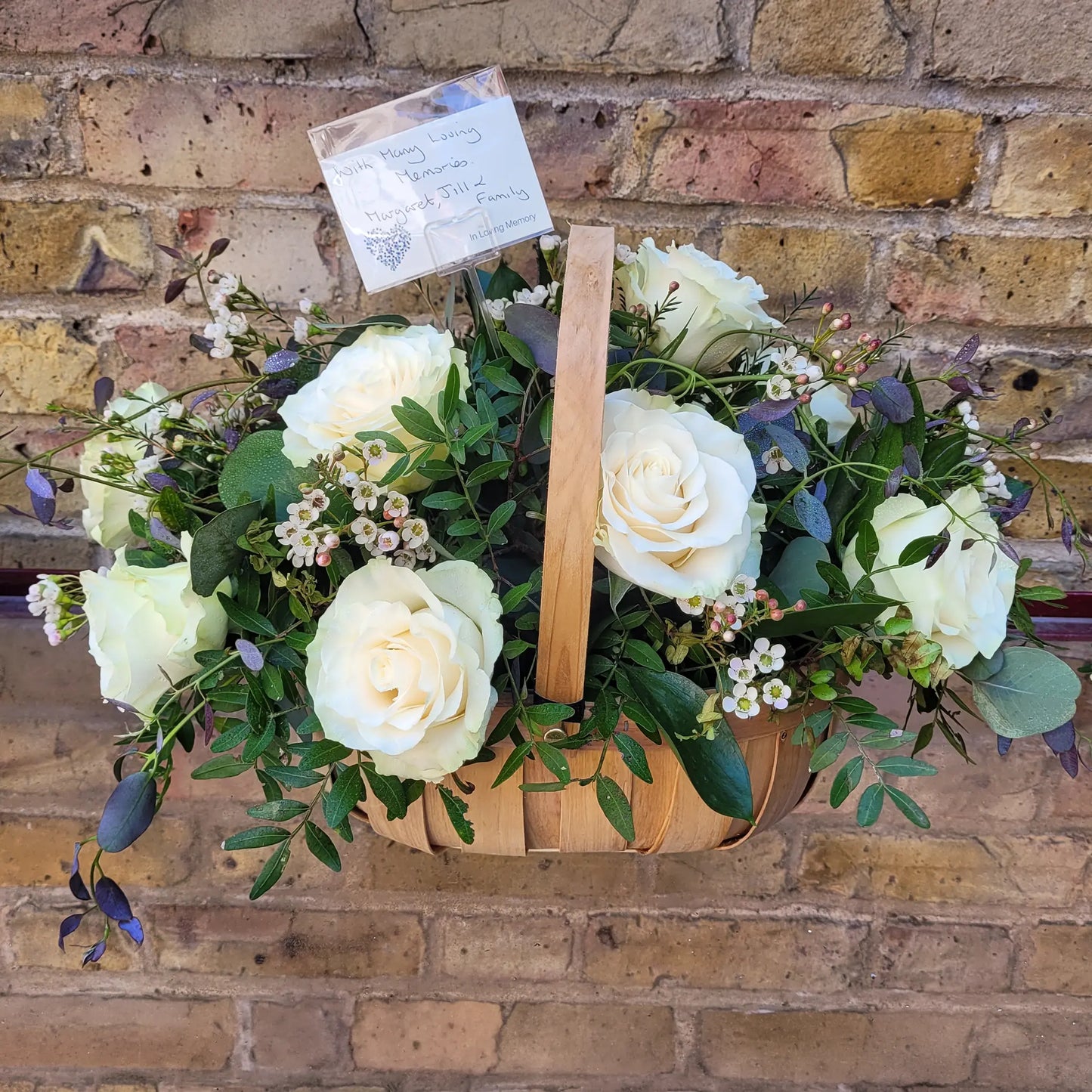 The ReCreation Flowers Funeral Flower Basket features florist choice flowers, white roses, and mixed foliage in a wooden basket with a small card, arranged in front of a rustic brick wall.