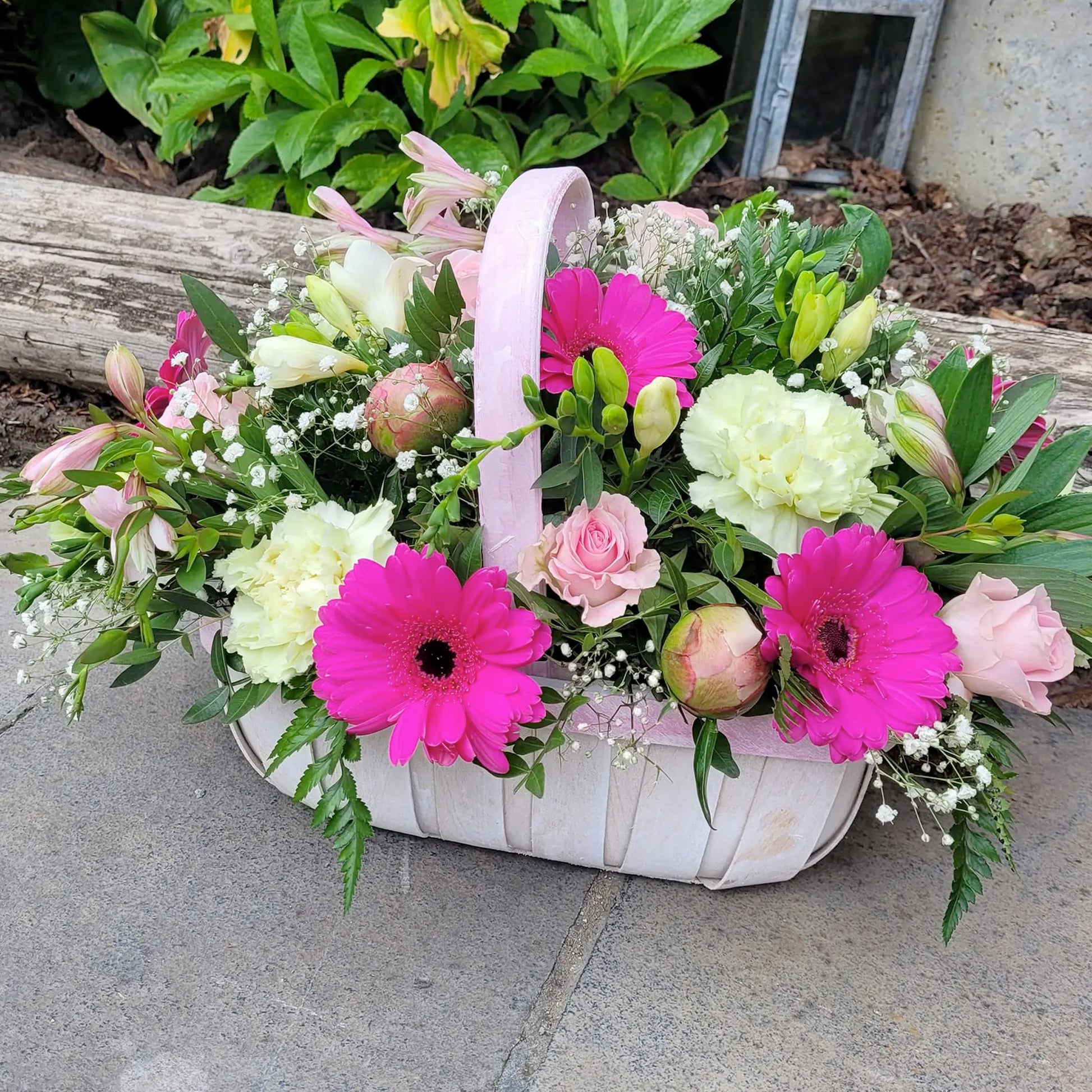 The Funeral Flower Basket by ReCreation Flowers features bright pink gerbera daisies, pale yellow carnations, light pink roses, and greenery in a white basket, displayed outdoors on stone.