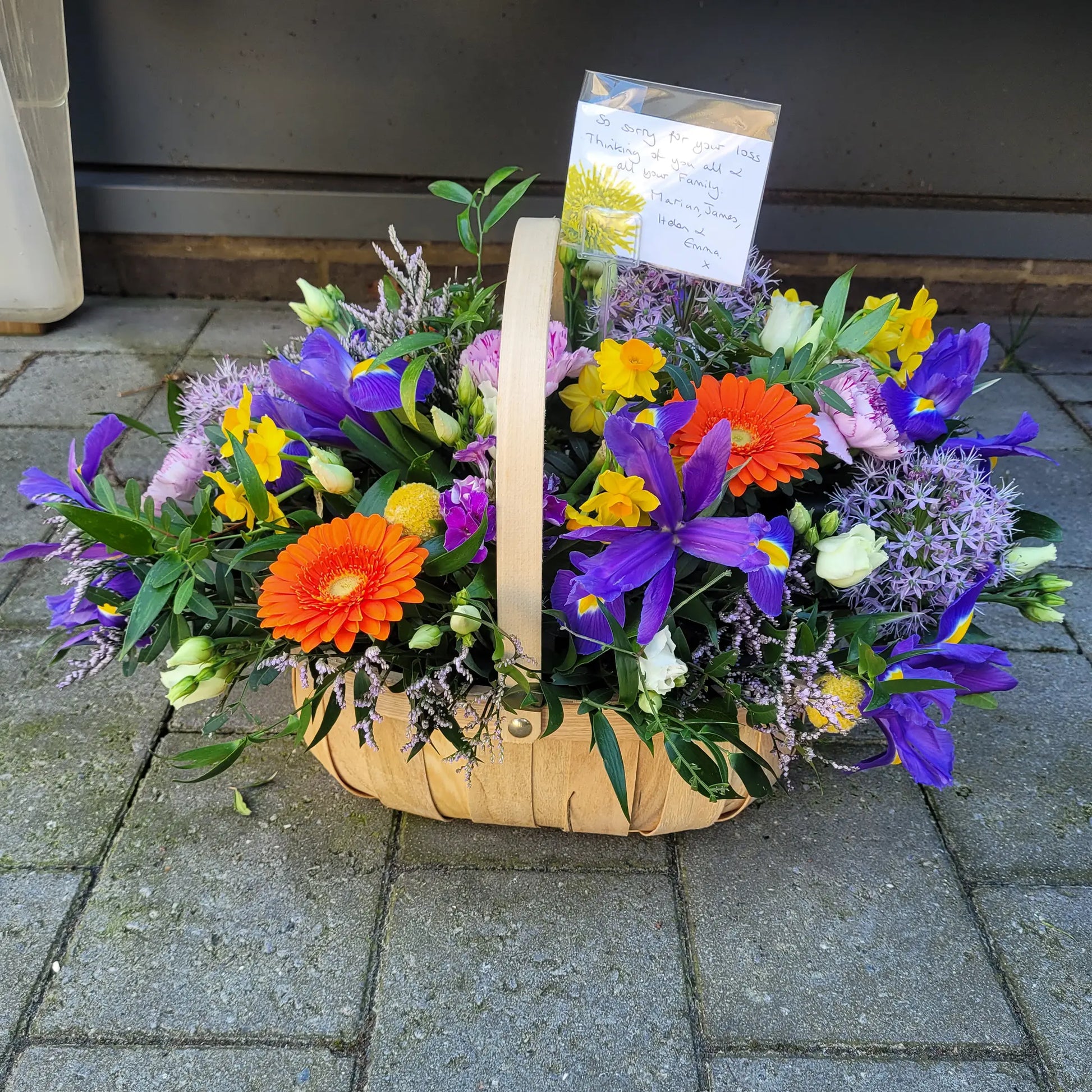 The Funeral Flower Basket by ReCreation Flowers features vibrant orange, purple, yellow, and white blooms arranged seasonally. A handwritten note on white paper rests among the florist’s choice flowers in the basket.