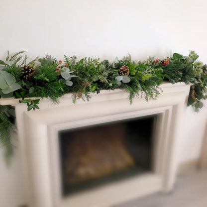 Get inspired for your next Christmas Garland Class with ReCreation Flowers: a white fireplace mantel adorned with a festive garland featuring fresh pine branches, eucalyptus leaves, pinecones, and red berries against a simple light-colored wall—the perfect seasonal touch for your holiday workshop.