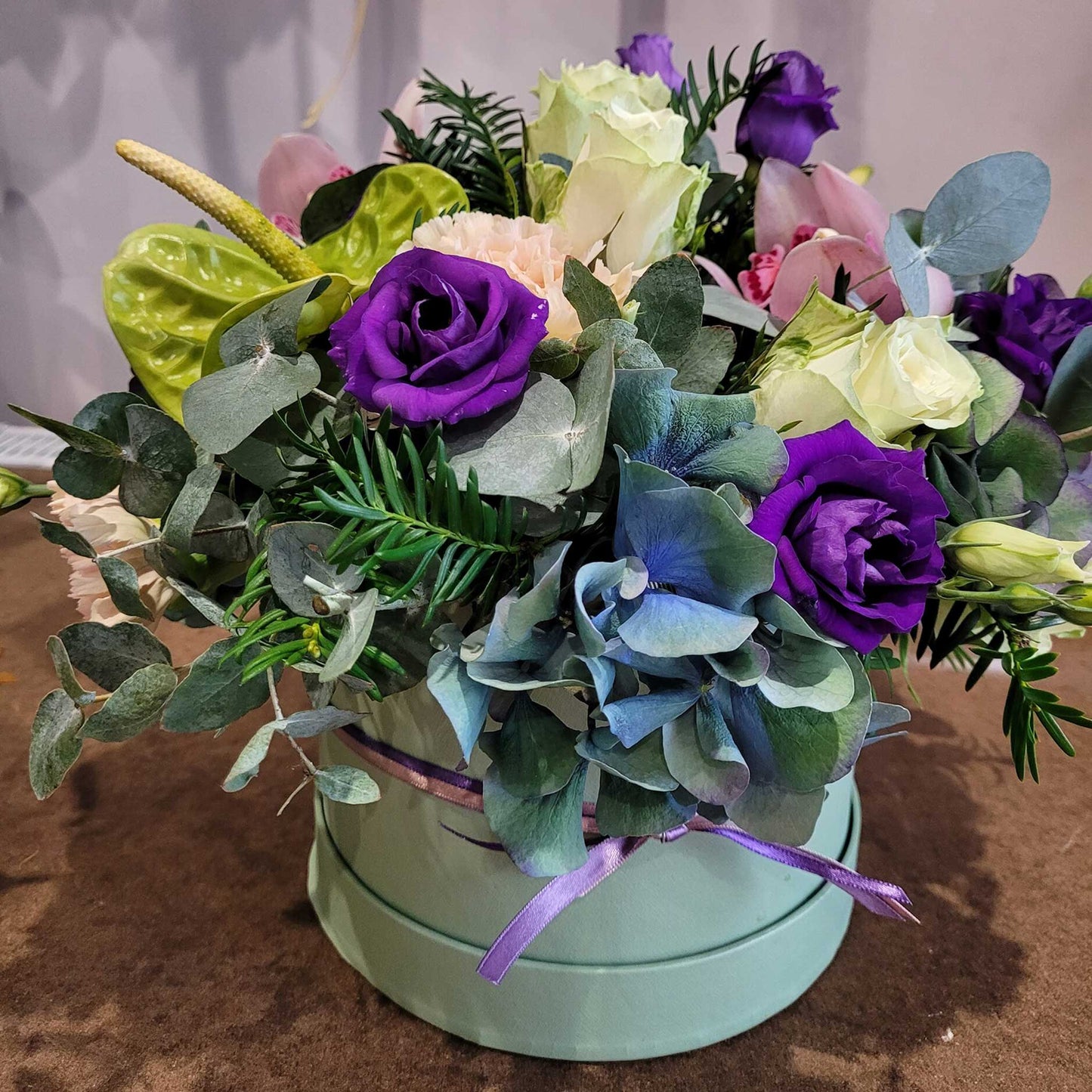 ReCreation Flowers’ Hatbox of Flowers features premium purple roses, blue hydrangeas, white and pink roses, and lush greenery in a mint-green hatbox with a purple ribbon—perfect for a lasting display.