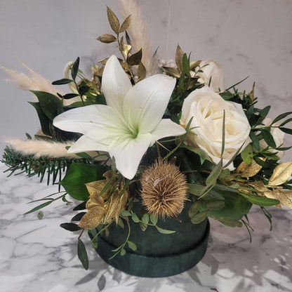 ReCreation Flowers’ Hatbox of Flowers showcases a large white lily, white rose, gold leaves, foliage, dried grasses, and a round dried flower in a dark green container on marble.