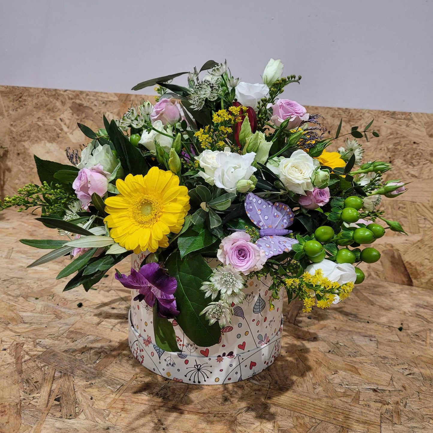 The ReCreation Flowers Hatbox of Flowers features yellow, pink, and white blooms with green foliage and a purple butterfly in a decorative round box, displayed on wood against a plain background.