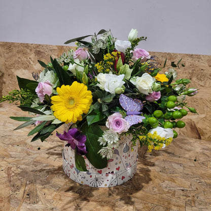 The ReCreation Flowers Hatbox of Flowers features yellow, pink, and white blooms with green foliage and a purple butterfly in a decorative round box, displayed on wood against a plain background.