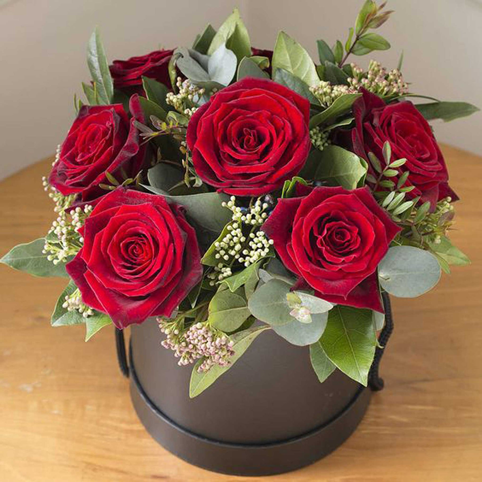 ReCreation Flowers' Hatbox of Flowers features vibrant red roses, lush green leaves, and delicate white accents beautifully arranged in a premium hatbox and displayed on a wooden table.