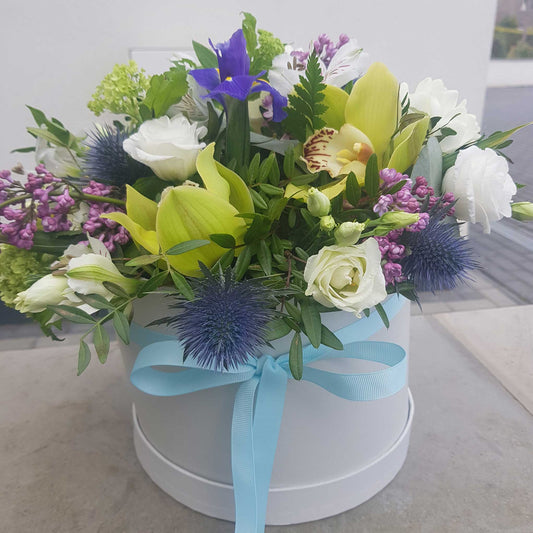 ReCreation Flowers' Hatbox of Flowers features premium yellow orchids, white roses, purple and blue blossoms, and green leaves, tied with a light blue ribbon—an elegant and long-lasting arrangement.