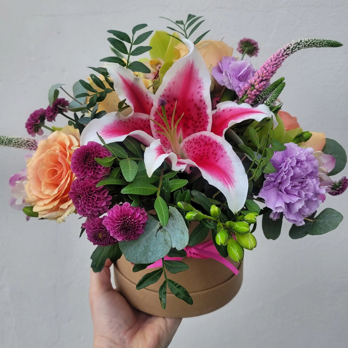 A hand holds the ReCreation Flowers Hatbox of Flowers, featuring pink lilies, peach roses, purple carnations, magenta accents, green leaves, and eucalyptus—artfully arranged for lasting beauty against a plain light background.