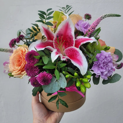A hand holds the ReCreation Flowers Hatbox of Flowers, featuring pink lilies, peach roses, purple carnations, magenta accents, green leaves, and eucalyptus—artfully arranged for lasting beauty against a plain light background.