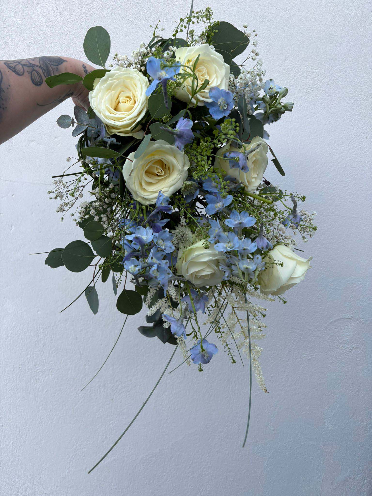 A hand holds a bouquet of cream-colored roses, blue delphiniums, babys breath, and green foliage against a plain light-gray background.