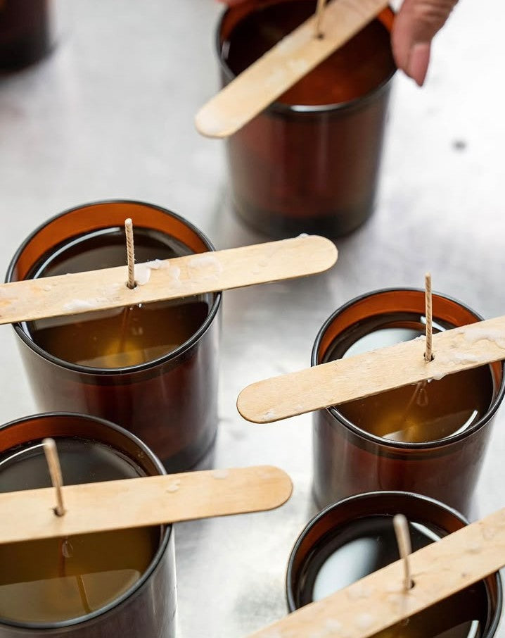A hand hovers over brown glass jars filled with wax and wooden sticks holding up candle wicks, illustrating the cozy process of candle making—a perfect activity for the Christmas Workshop @ GAS by ReCreation Flowers or a festive wreath making session at home.