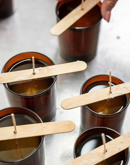 A hand hovers over brown glass jars filled with wax and wooden sticks holding up candle wicks, illustrating the cozy process of candle making—a perfect activity for the Christmas Workshop @ GAS by ReCreation Flowers or a festive wreath making session at home.
