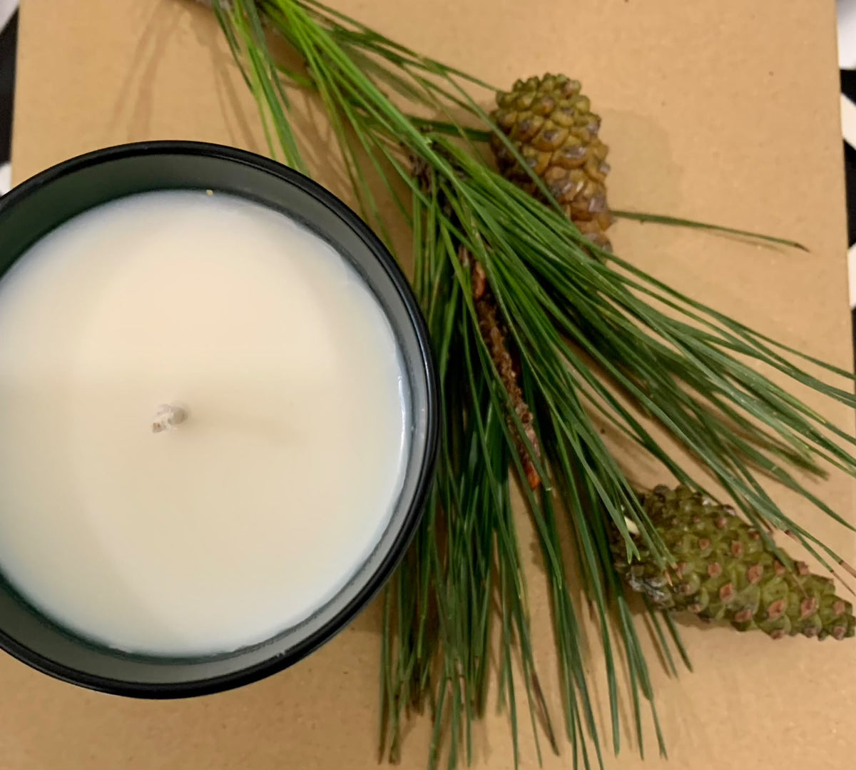A “Christmas Workshop @ GAS” candle by ReCreation Flowers, in a white wax and sleek black container, rests beside green pine needles and three small pinecones on a brown surface—capturing the festive charm of wreath-making and holiday creativity.
