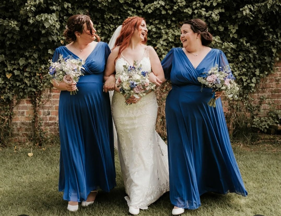 Three women stand arm in arm, smiling and laughing. The woman in the middle wears a white wedding dress and veil, while the two women on either side wear matching blue dresses and hold bouquets. They are outdoors by a brick wall with greenery.