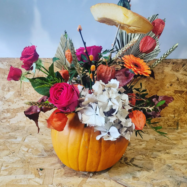 A pumpkin serves as a vase for a vibrant floral arrangement featuring roses, dried leaves, orange blooms, and a decorative slice of pumpkin on top. Offered by ReCreation Flowers, the Autumn Flower Class in Leigh-on-Sea lets you create this stunning seasonal display—the perfect centerpiece for your autumn workshop table.