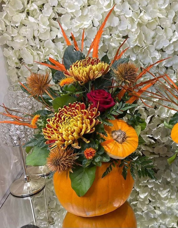 A floral arrangement created at the Autumn Flower Class by ReCreation Flowers in Leigh-on-Sea features yellow and red blooms, orange accents, and greenery artfully arranged in a hollowed pumpkin. The design is complemented by a white hydrangea backdrop and accompanied by a glass lamp.