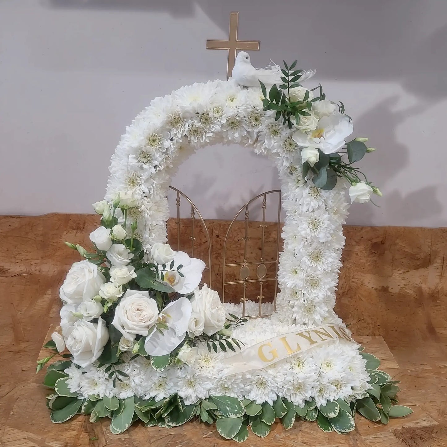 The Special Tributes by ReCreation Flowers features an arch-shaped floral arrangement with white flowers, a gold gate center, a cross on top, white dove, "GLYN" ribbon, and green leaves and white roses at the base. A unique custom tribute design.