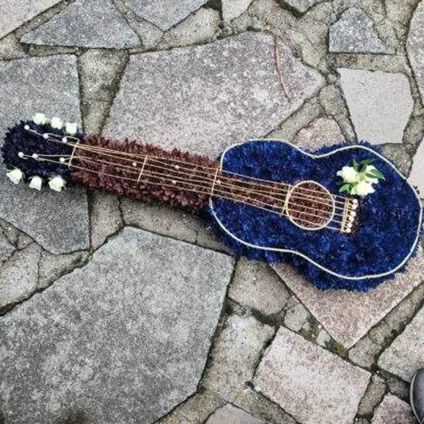 A unique Special Tributes arrangement by ReCreation Flowers, this guitar-shaped floral tribute features blue and brown blooms with white accents and string details, artfully displayed on a stone pavement.