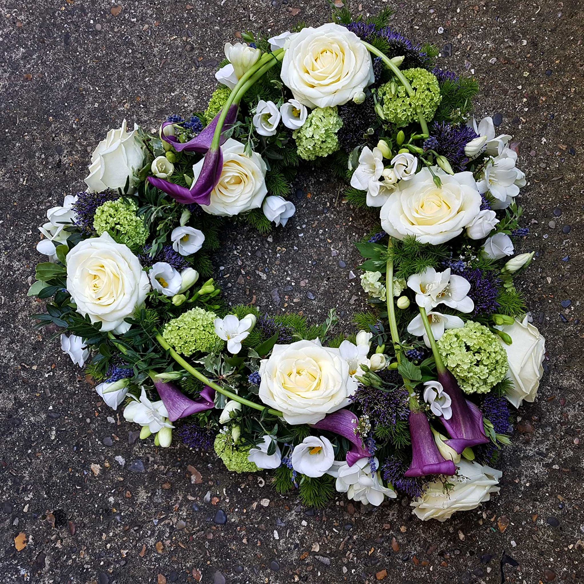 The Open Heart Funeral Flowers - Loose Style by ReCreation Flowers features a heart-shaped wreath with white roses, green hydrangeas, purple calla lilies, and small white blossoms as a graceful funeral tribute.