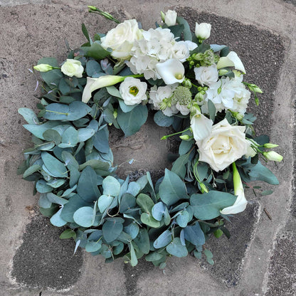 The Funeral Flower Wreath by ReCreation Flowers features white roses, calla lilies, and silver-green eucalyptus leaves—a timeless floral tribute resting elegantly, perfect for funeral arrangements.