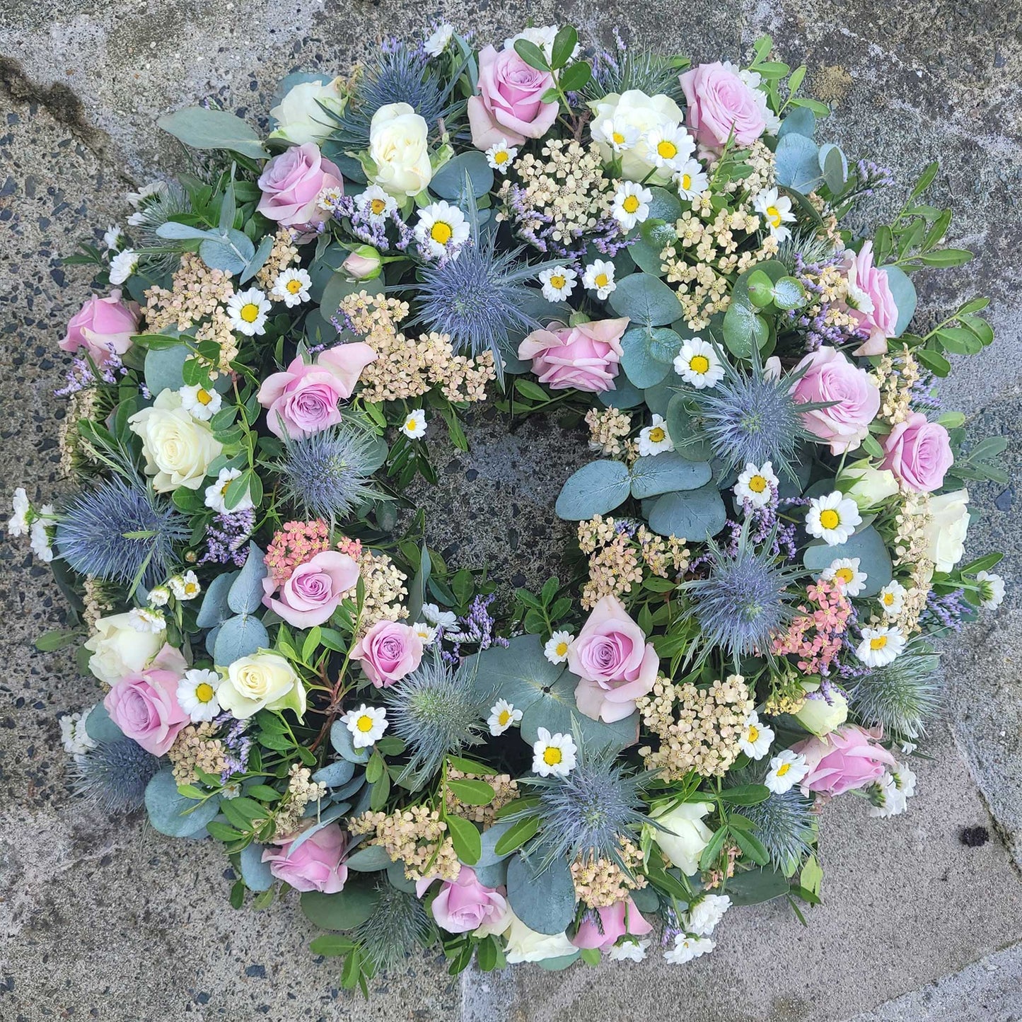 The Funeral Flower Wreath by ReCreation Flowers features pink, white, and cream roses, daisies, blue thistles, pale yellow blooms, eucalyptus leaves, and greenery elegantly arranged for funerals on a textured stone surface.