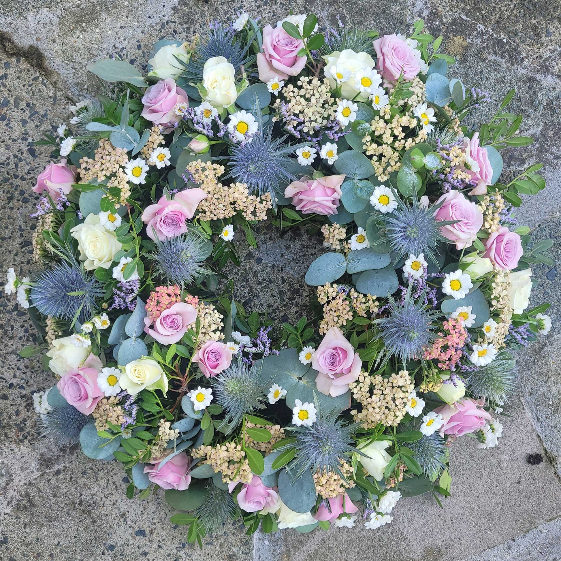 The Funeral Flower Wreath by ReCreation Flowers features pink, white, and cream roses, daisies, blue thistles, pale yellow blooms, eucalyptus leaves, and greenery elegantly arranged for funerals on a textured stone surface.