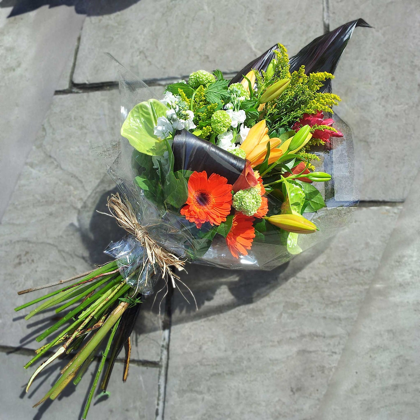 Funeral Sheaves by ReCreation Flowers: a beautiful seasonal bouquet of orange gerbera daisies, yellow lilies, green foliage, and dark leaves, tied with twine and wrapped in clear plastic—perfect for heartfelt tributes.