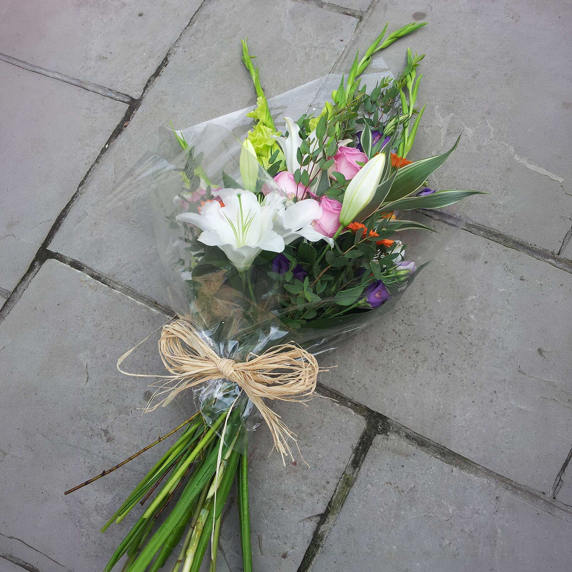 ReCreation Flowers' Funeral Sheaves, featuring white lilies and pink roses, wrapped in clear plastic with a raffia bow, rest on gray stone pavement.