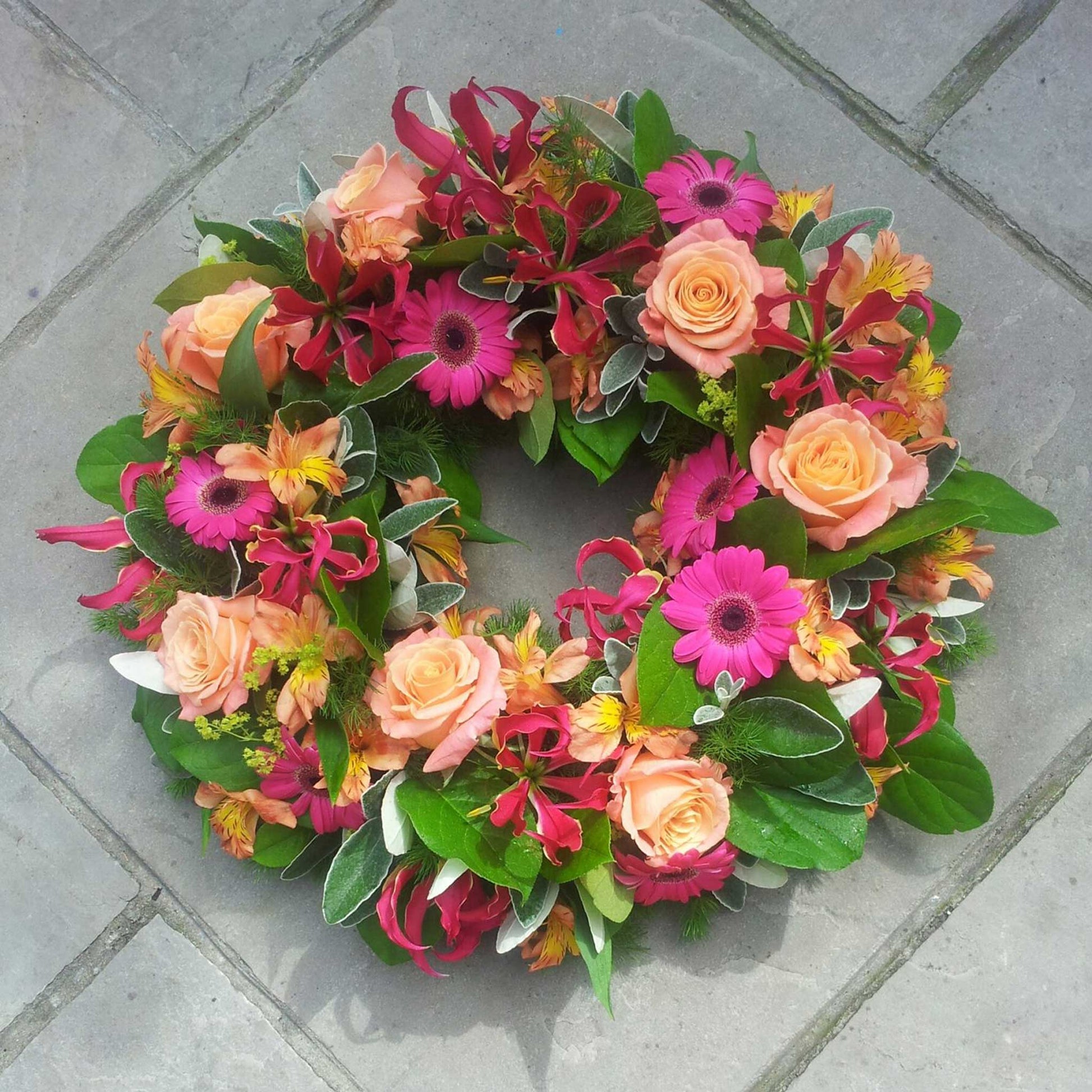 The Funeral Flower Wreath by ReCreation Flowers features peach roses, pink gerberas, yellow alstroemeria, red gloriosa lilies, and greenery—a stunning funeral arrangement displayed on a gray stone surface.