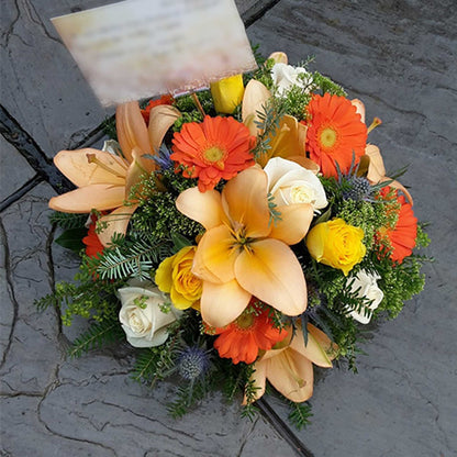 The Funeral Flower Posy by ReCreation Flowers features a compact round arrangement of vibrant seasonal blooms, including orange lilies, orange gerberas, yellow and white roses, and greenery—beautifully displayed on a stone surface.
