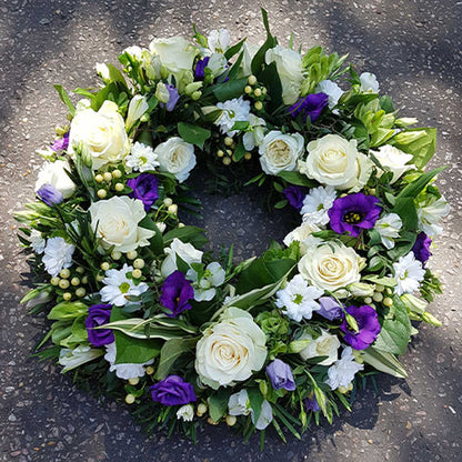 The Funeral Flower Wreath by ReCreation Flowers features white roses, purple blooms, small white petals, and lush greenery on a textured gray base—a tasteful arrangement ideal as funeral flowers.