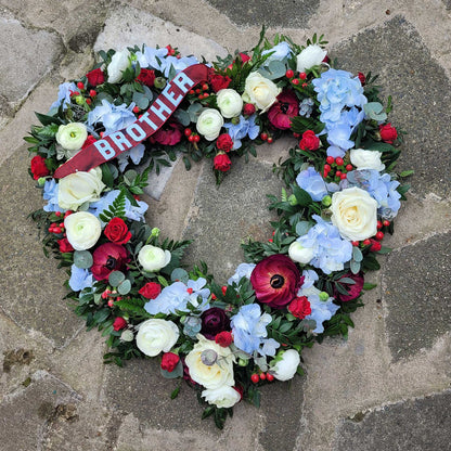 The Open Heart Funeral Flowers - Loose Style by ReCreation Flowers features an open heart design with white, red, and blue blooms, green foliage, and a red BROTHER ribbon, delicately arranged on a stone pavement.