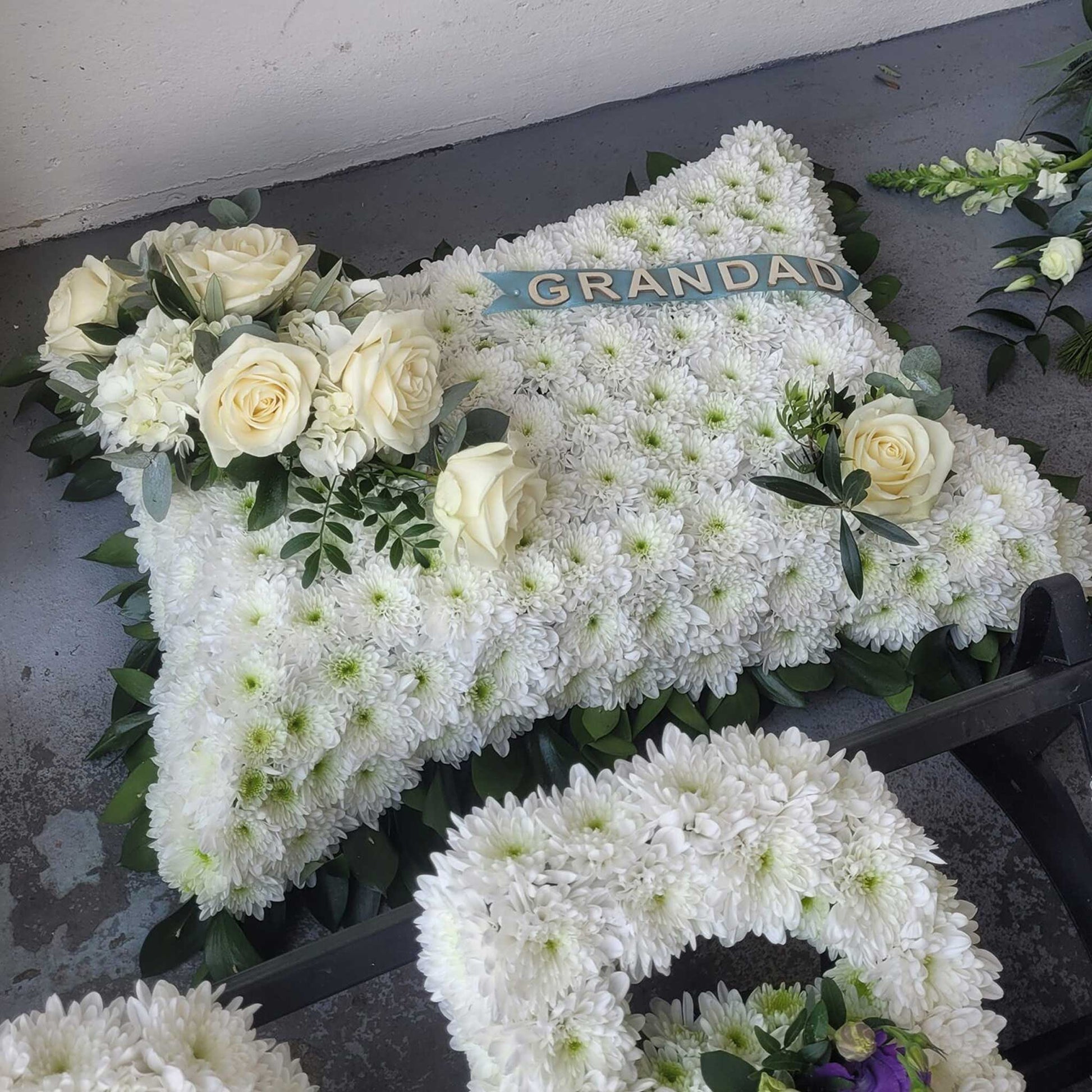 A ReCreation Flowers "Cushions & Pillows - White Base" funeral pillow with white roses, greenery, and a GRANDAD ribbon is shown on a gray surface beside part of a white flower wreath.