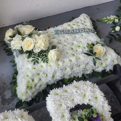 A ReCreation Flowers "Cushions & Pillows - White Base" funeral pillow with white roses, greenery, and a GRANDAD ribbon is shown on a gray surface beside part of a white flower wreath.