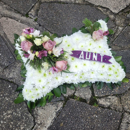 A Cushions & Pillows - White Base tribute by ReCreation Flowers, featuring pink and purple seasonal blooms and a "AUNT" ribbon, is thoughtfully placed on a stone pavement.