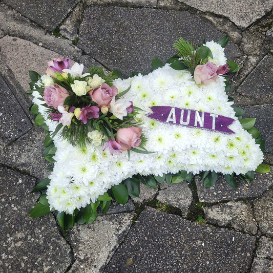 A Cushions & Pillows - White Base tribute by ReCreation Flowers, featuring pink and purple seasonal blooms and a "AUNT" ribbon, is thoughtfully placed on a stone pavement.