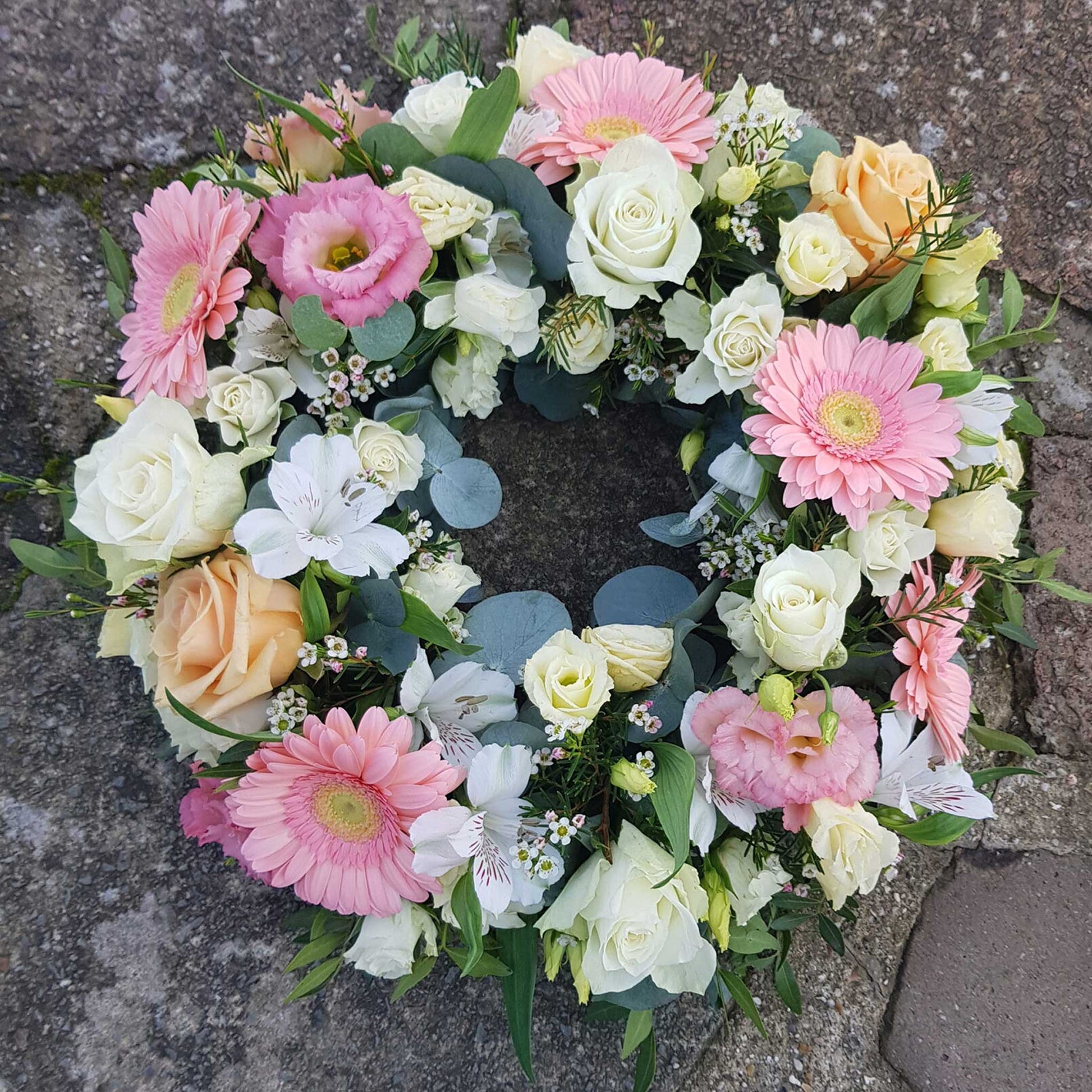 The Funeral Flower Wreath by ReCreation Flowers features pink gerbera daisies, white and pale peach roses, lilies, and greenery in a circular arrangement, offering an elegant tribute displayed on a stone surface.
