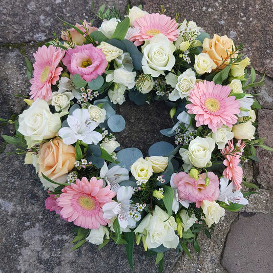 The Funeral Flower Wreath by ReCreation Flowers features pink gerbera daisies, white and pale peach roses, lilies, and greenery in a circular arrangement, offering an elegant tribute displayed on a stone surface.