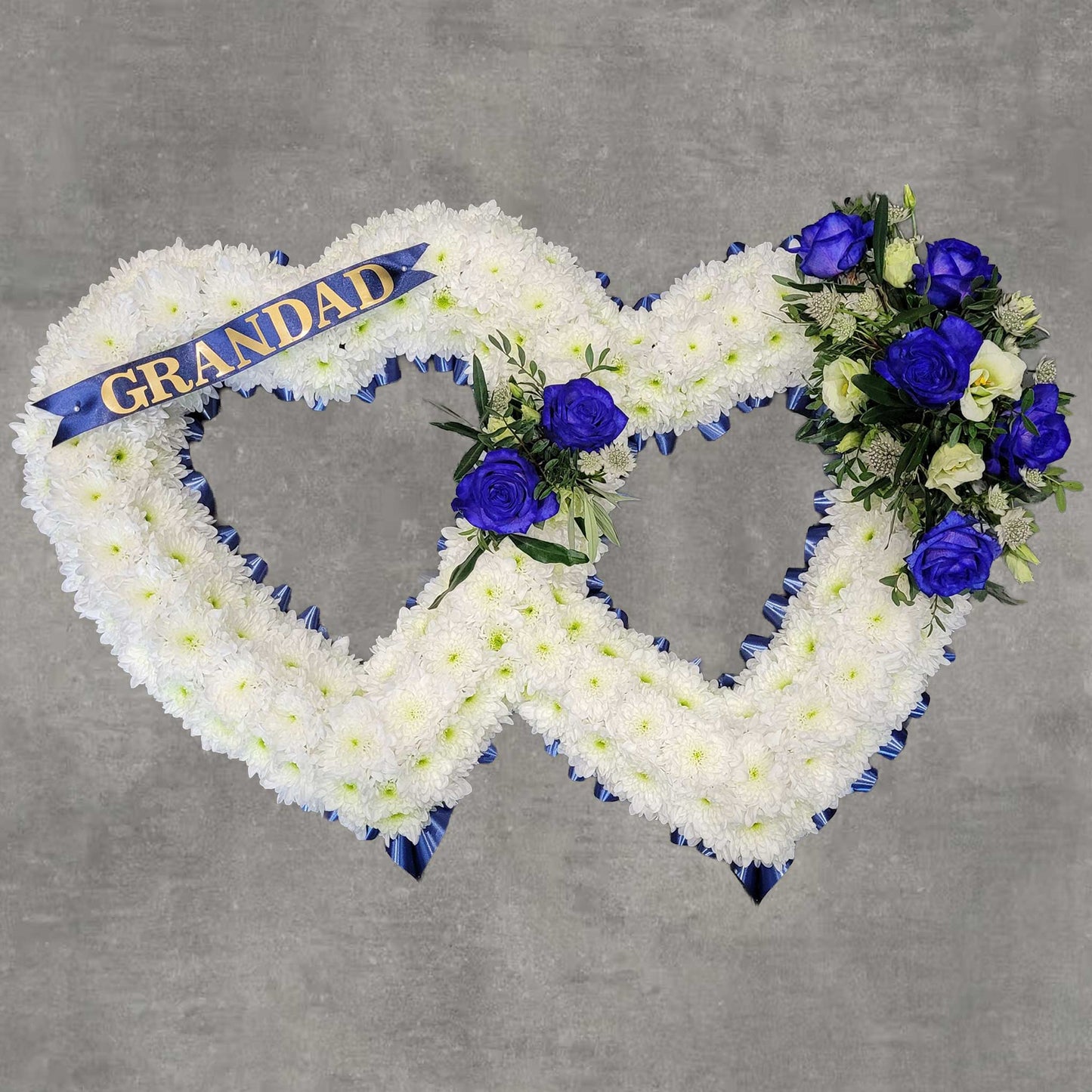 The Open Heart Funeral Flowers by ReCreation Flowers features two interlocking white floral hearts, adorned with blue and purple roses, seasonal blooms, greenery, and a blue "GRANDAD" ribbon on a gray background.