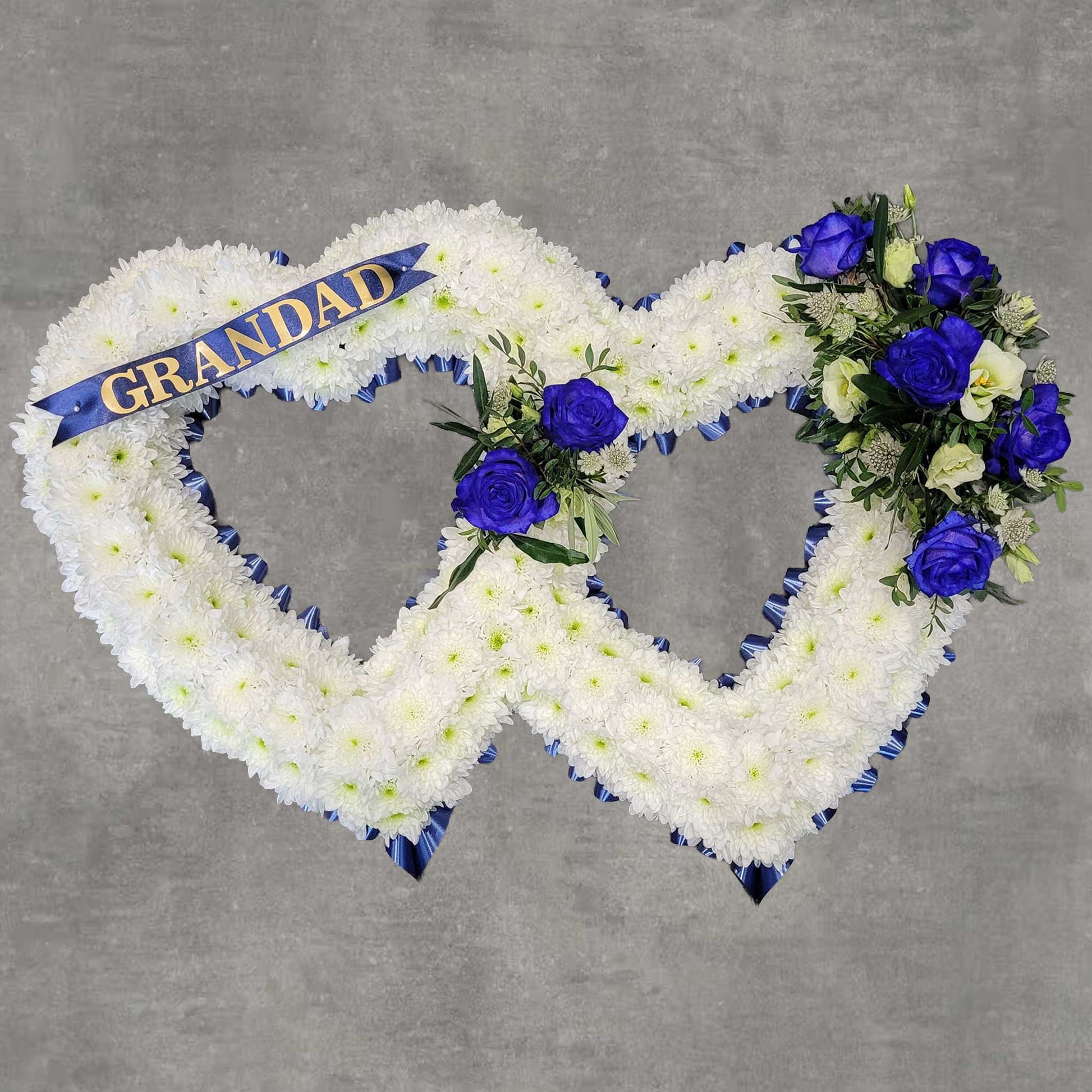 The Open Heart Funeral Flowers by ReCreation Flowers features two interlocking white floral hearts, adorned with blue and purple roses, seasonal blooms, greenery, and a blue "GRANDAD" ribbon on a gray background.